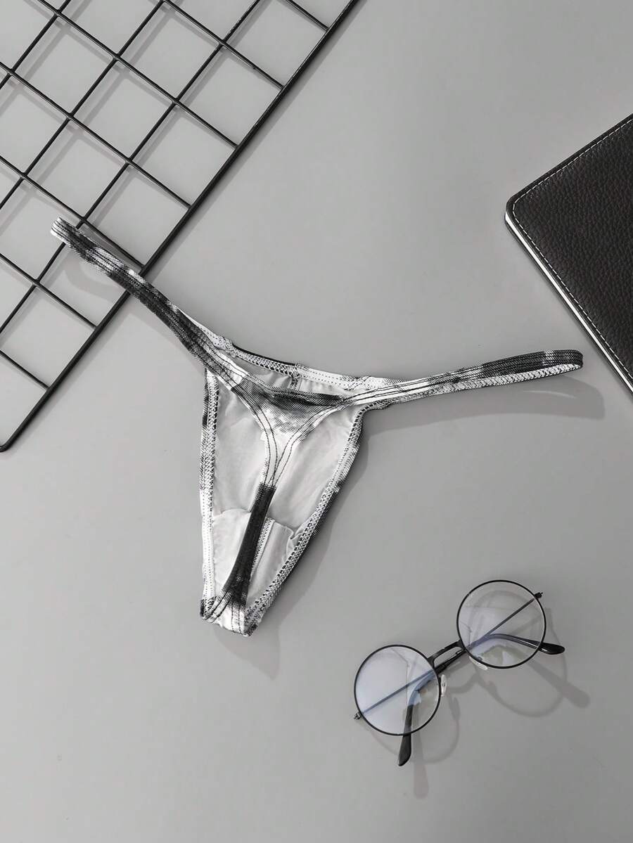 Black men's low rise thong underwear featuring asymmetrical tie dye design made from knitted polyester fabric.